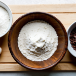 A meticulously arranged flat lay of premium brownie baking ingredients on a clean, light-colored wooden countertop. Focus on a pile of dark chocolate callets (70% cacao), a stick of unsalted butter, a bowl of light brown sugar, a bowl of white granulated sugar, a few fresh eggs in a rustic ceramic bowl, a small sifter with all-purpose flour, and a sprinkle of sea salt. The scene is well-lit with soft, natural light, emphasizing texture and quality. The overall aesthetic is clean, professional food photography. perfect anatomy, correct proportions, natural pose, well-formed hands (if any are visible, though the focus is on ingredients), natural body proportions, safe for work, appropriate content, fully clothed, professional.